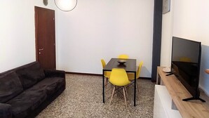 Un espacio habitable pequeño con un sofá de color oscuro, una mesa de comedor con sillas amarillas, un televisor sobre un mueble de madera y una lámpara colgante.