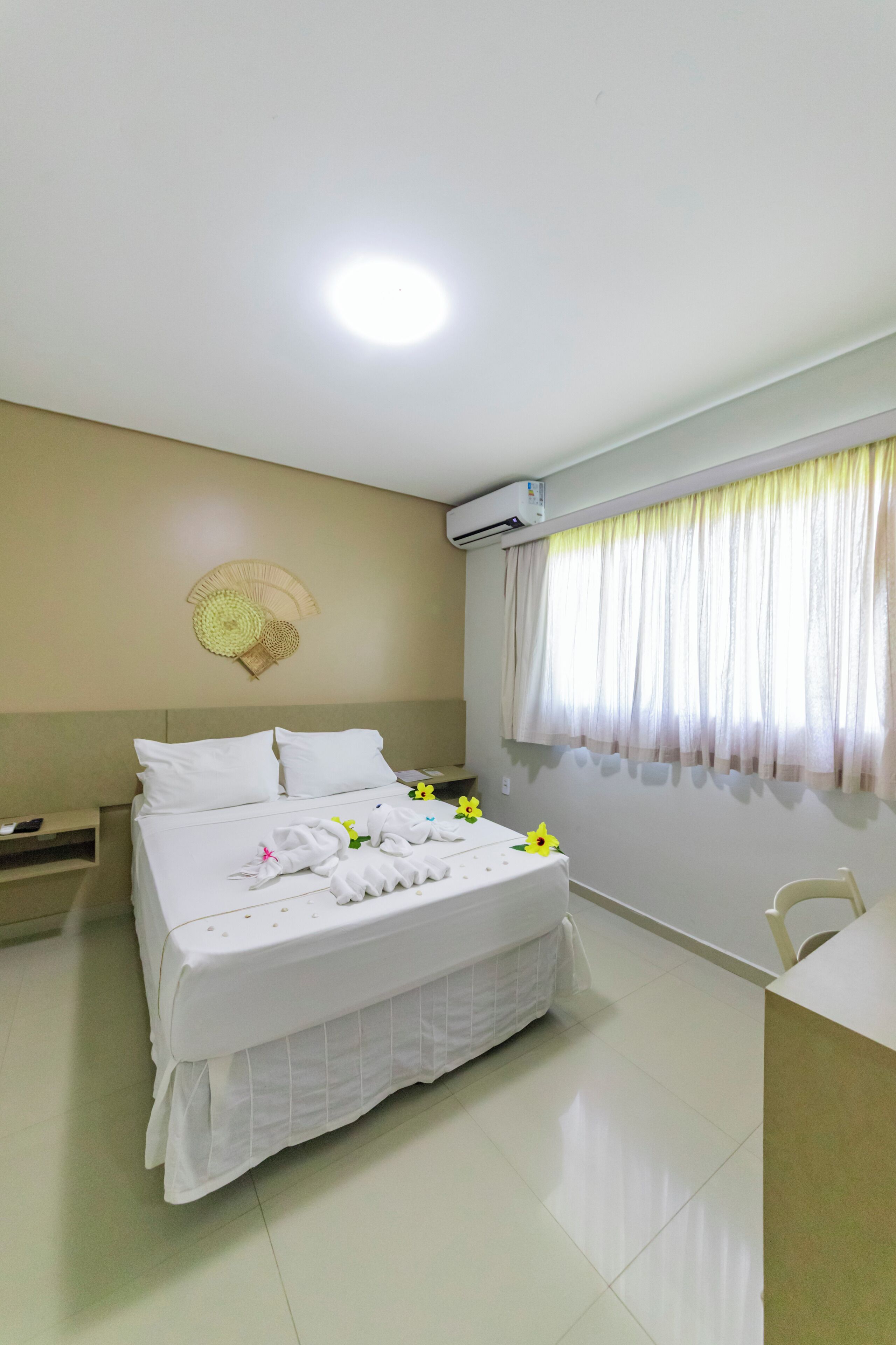 A neatly made bed with white linens, a bedside table, a chair, and a window with sheer curtains.