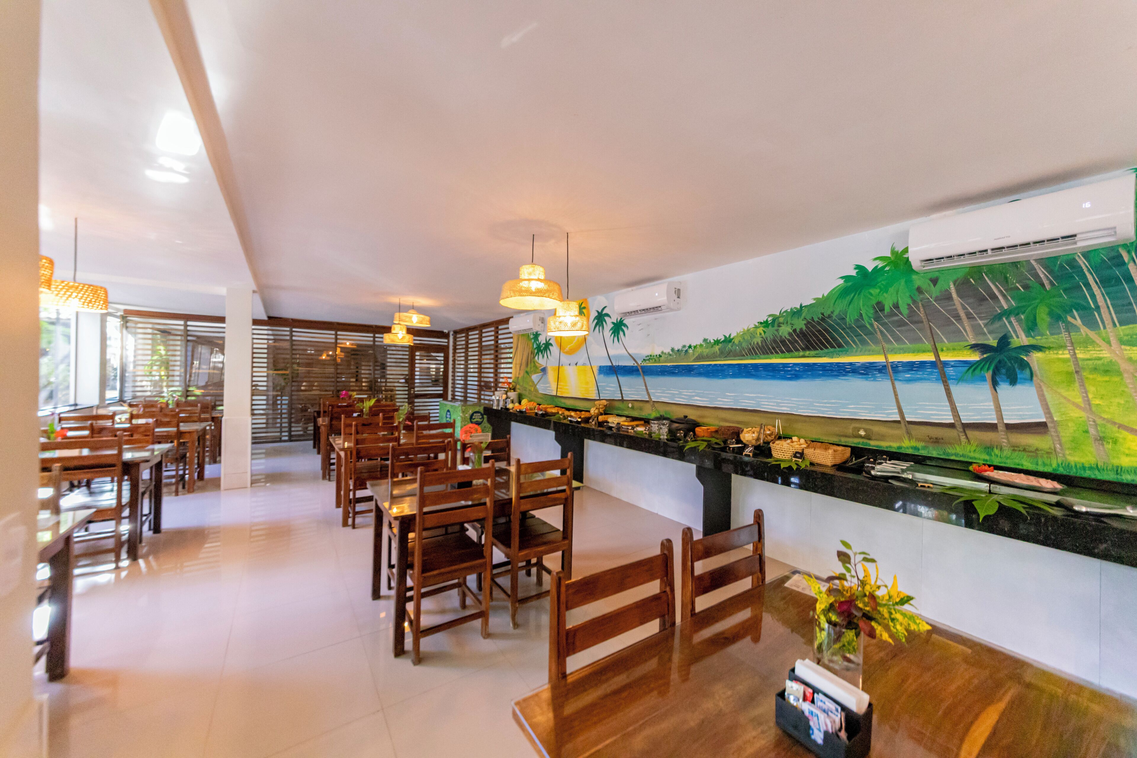 Restaurante com mesas e cadeiras de madeira, um balcão comprido com vista para a praia e palmeiras, e luminárias pendentes.