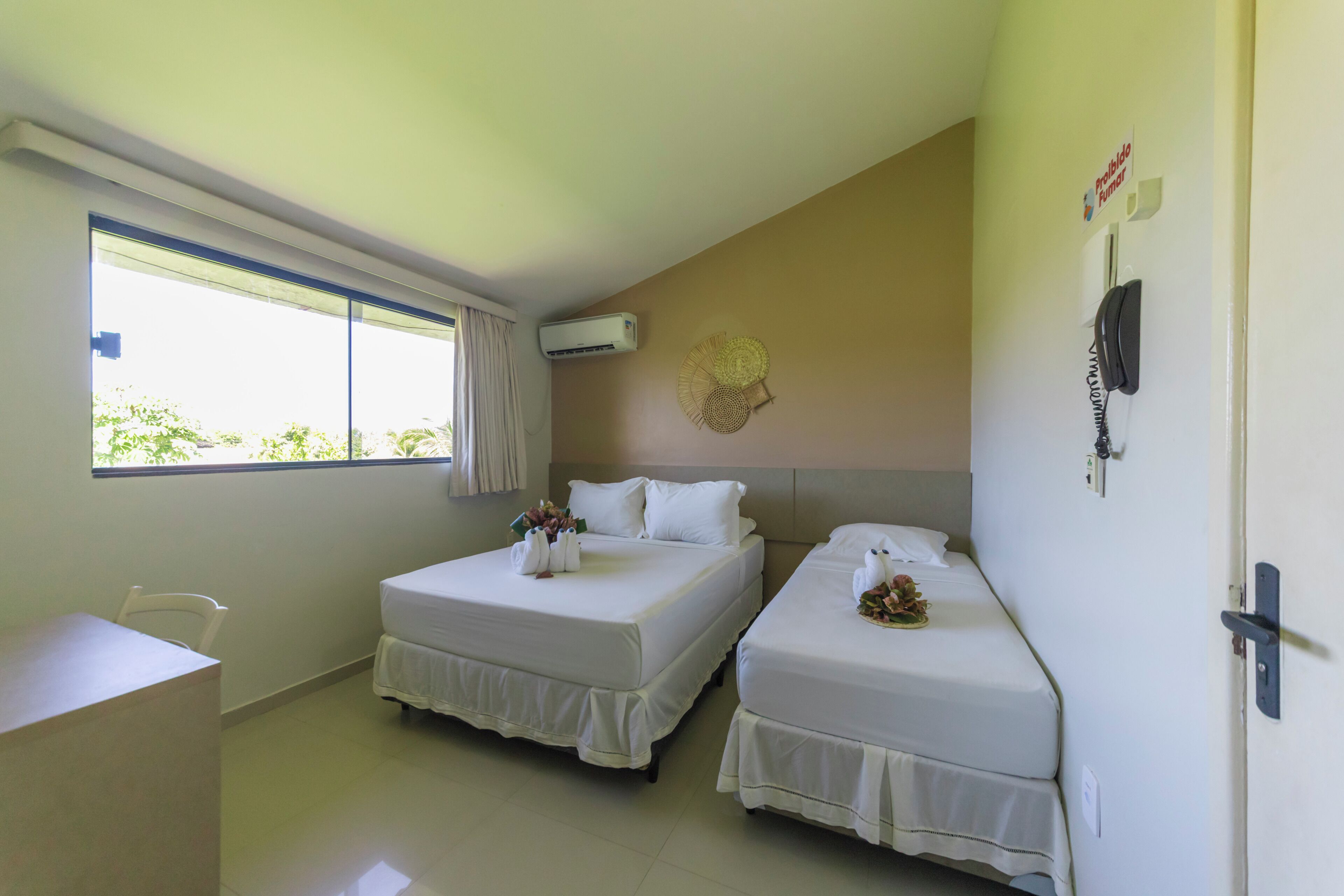 A hotel room with two beds, a desk, a chair, a window with curtains, an air conditioning unit, a wall-mounted phone, and a decorative wall piece.
