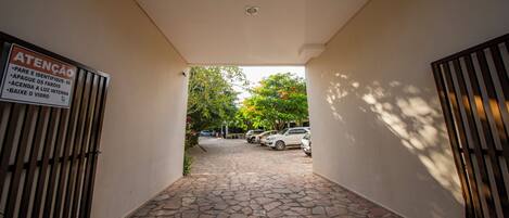 A stone-paved pathway leading to a parking area with cars, surrounded by trees and a signboard on the left.