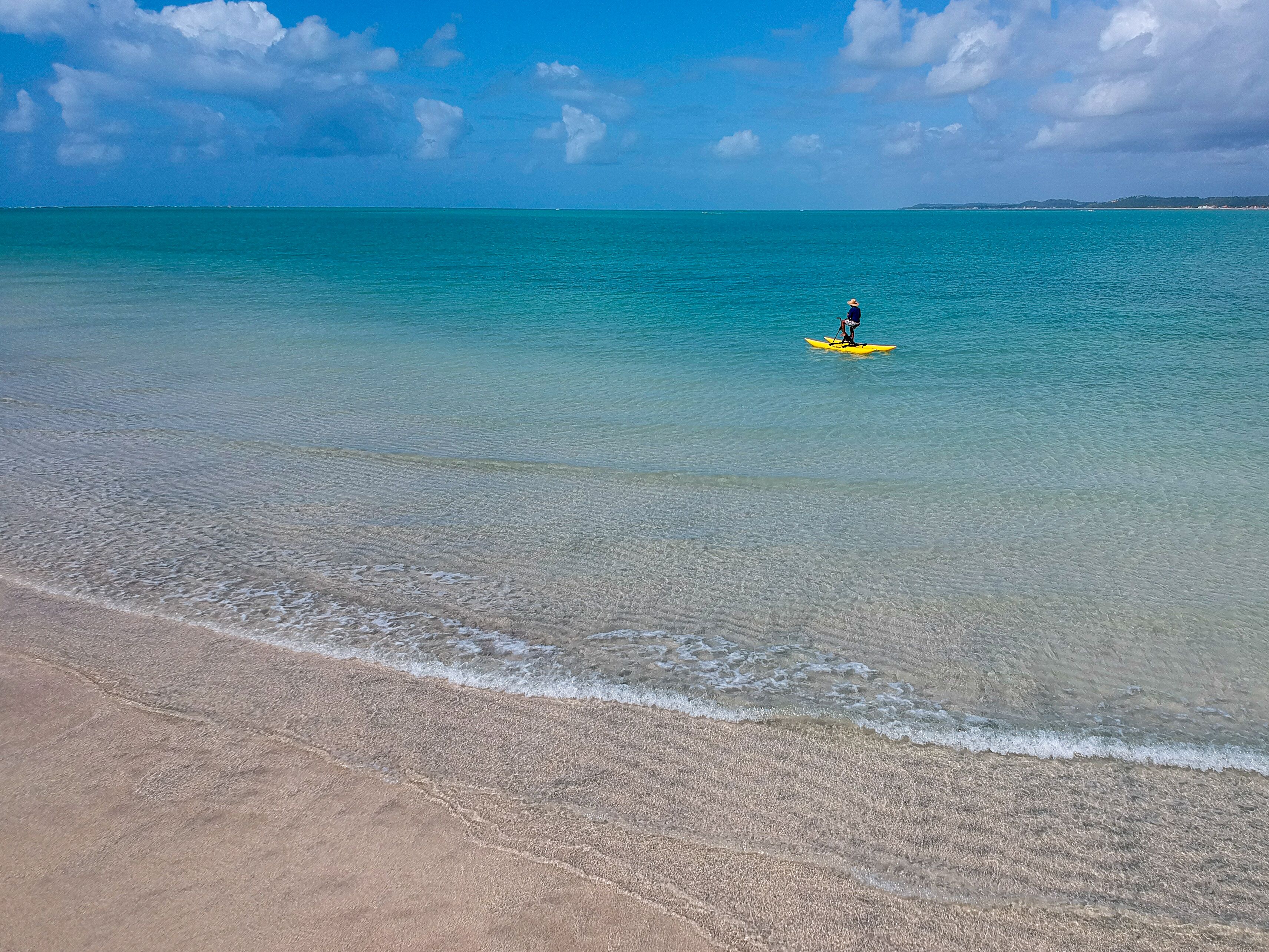 Une plage avec une eau turquoise et limpide, et une personne qui rame sur un kayak jaune.