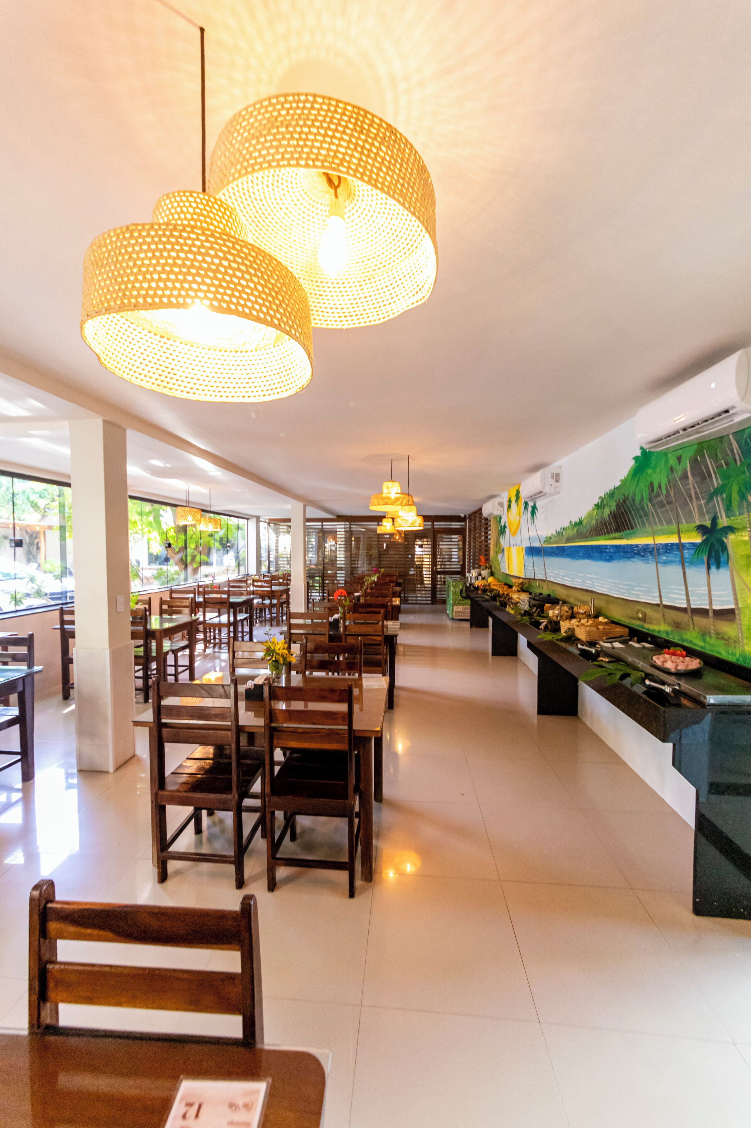 Un restaurante con mesas y sillas de madera, lámparas colgantes de tejido y un mural con una escena de playa tropical.