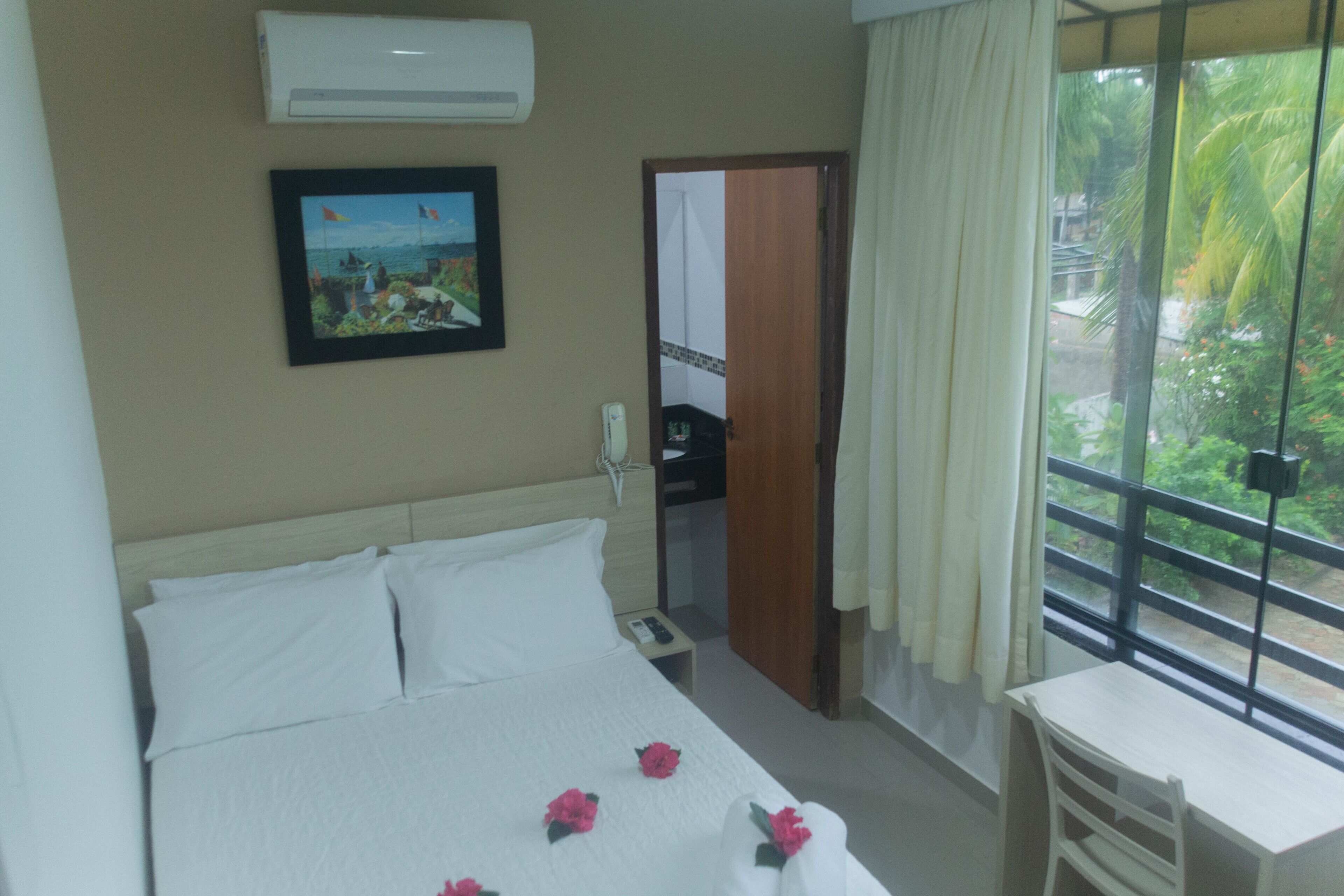 Habitación de hotel con cama, escritorio, ventana con vistas a zonas verdes, aire acondicionado y un cuadro en la pared.