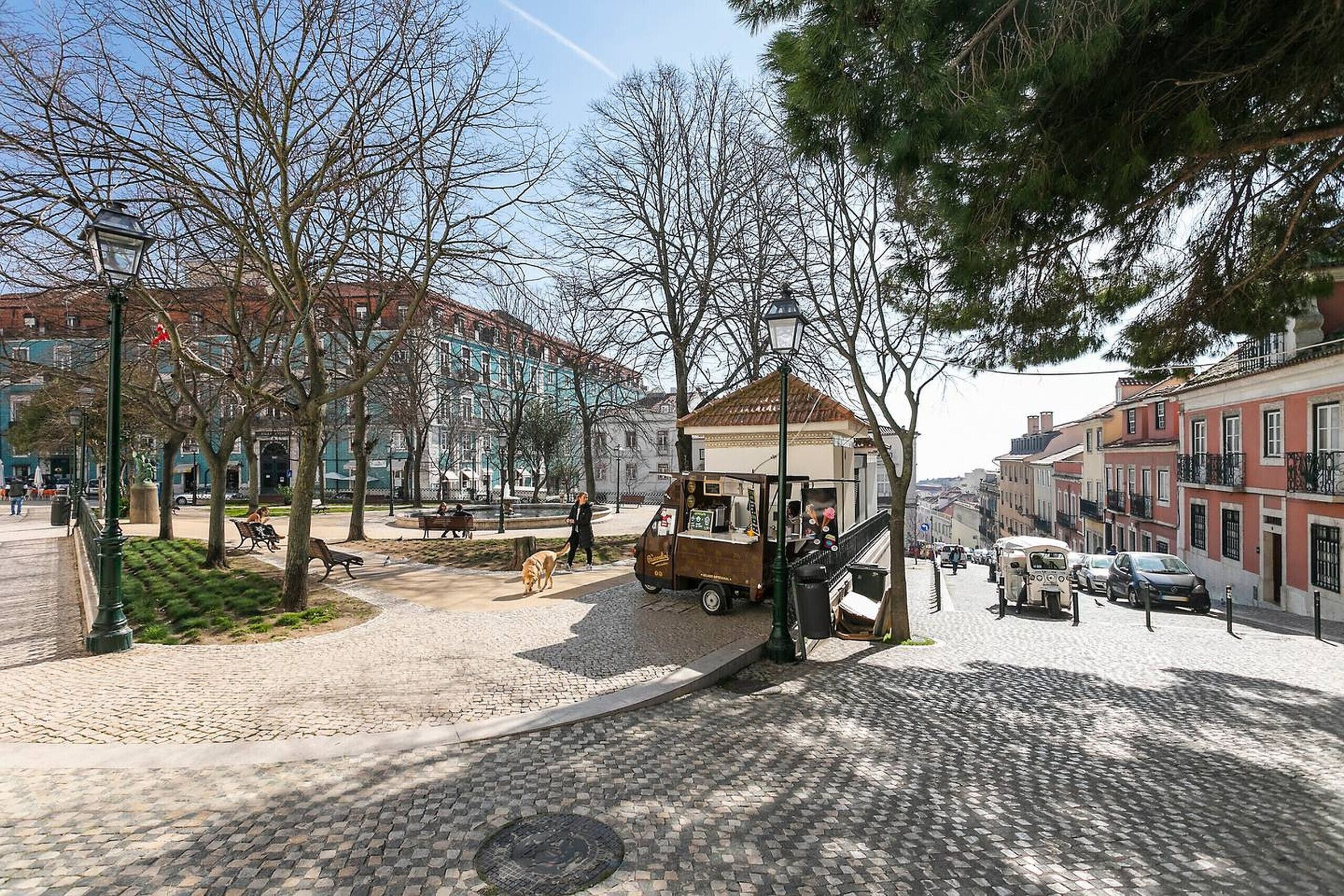 A cobblestone square with a food cart, benches, and a dog walking.