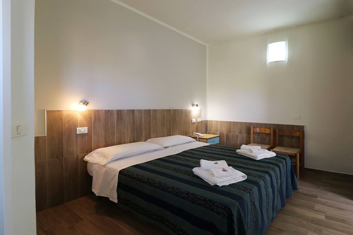 A hotel room with two single beds, wooden headboards, a small bedside table with a lamp, and a chair.