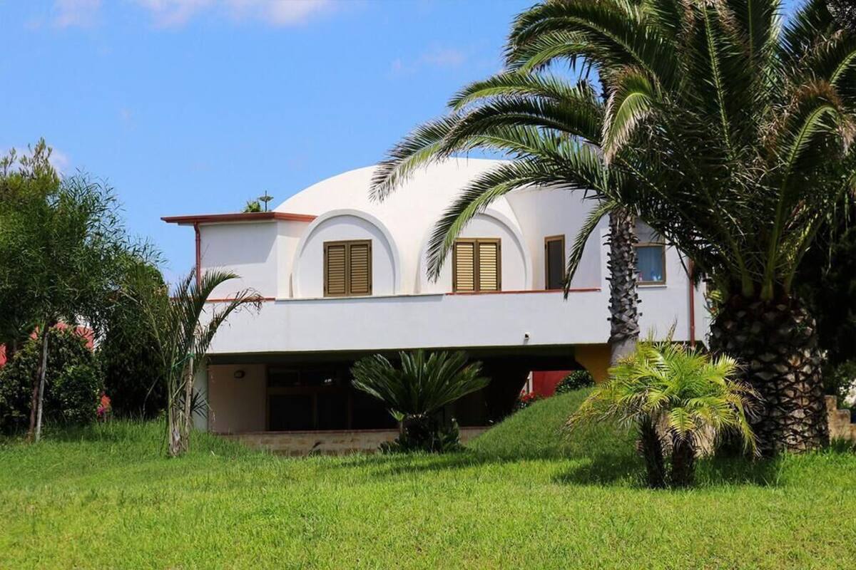 A white building with a curved roof and arched windows, surrounded by lush greenery and palm trees.