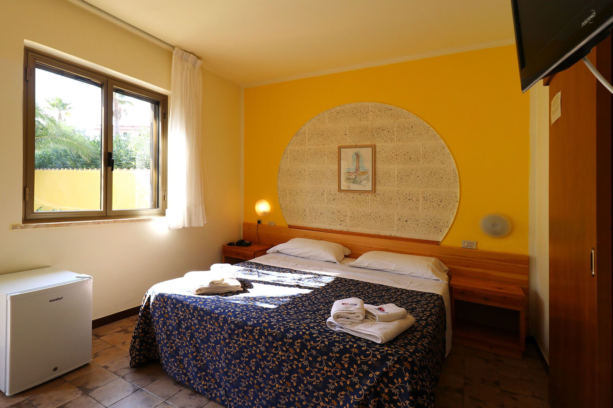 A hotel room with a bed, a small bedside table, a lamp, a heater, and a window with curtains.