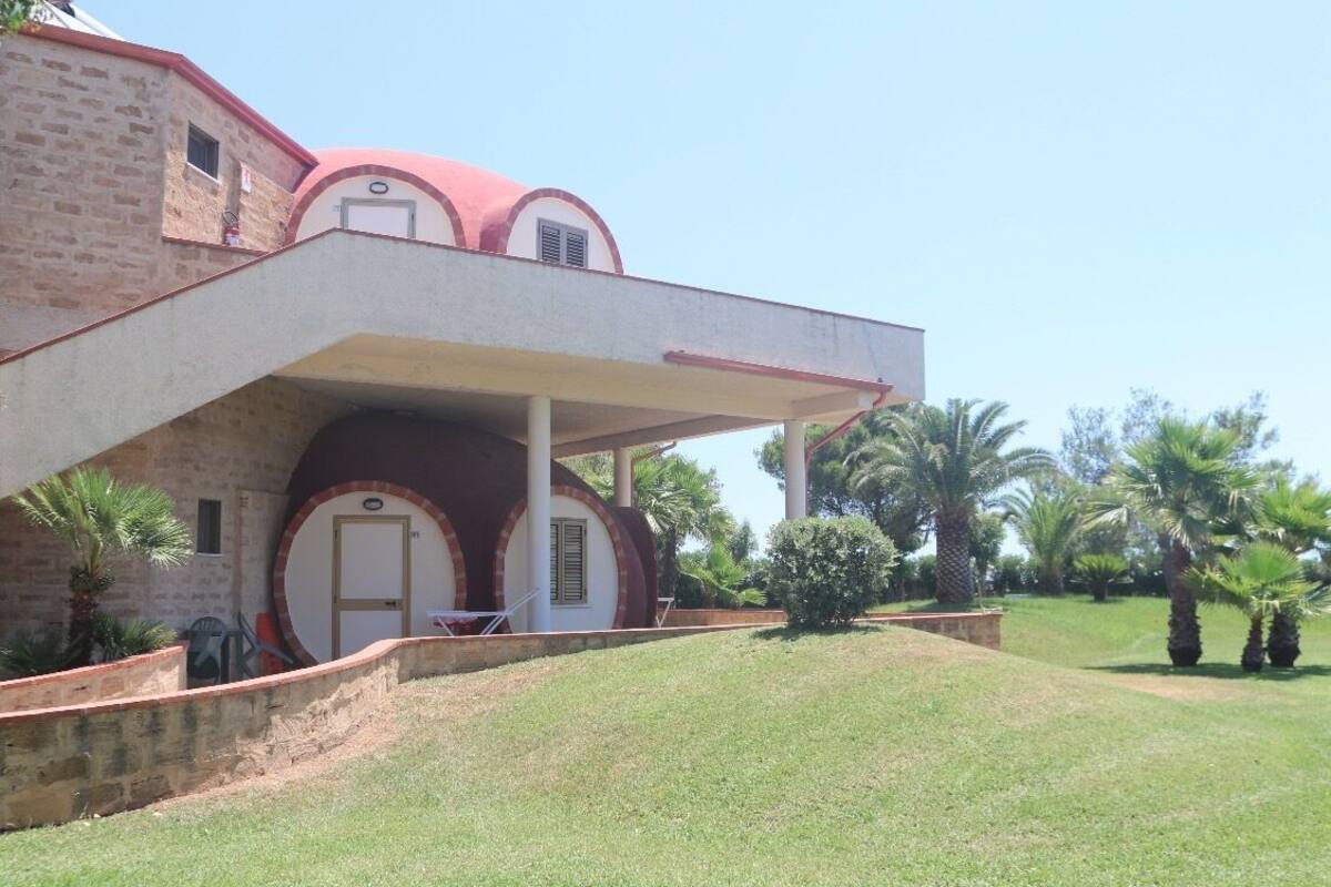 A two-story building with a red-tiled roof, a covered porch, and a small garden area with palm trees.