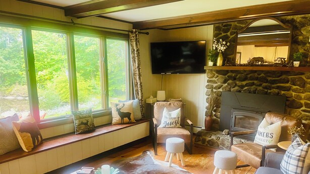 A cozy living room with a stone fireplace, a comfortable sofa, a chair with a 'Be Our Guest' pillow, a television, and a window with a view of greenery. - Perfect Lake or Mountain Getaway Less Than 5 Minutes Away (Sunapee)