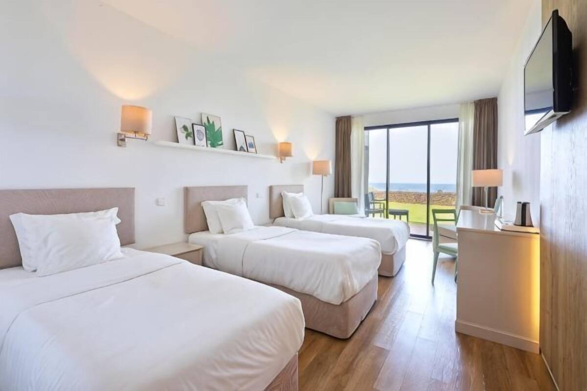 Ein Hotelzimmer mit drei Betten, einem Schreibtisch, einem Fernseher und einem großen Fenster mit Blick.
