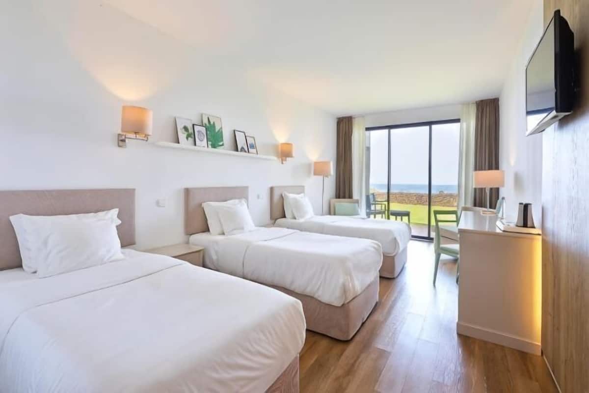 A hotel room with three beds, a desk, a TV, and a large window with a view.
