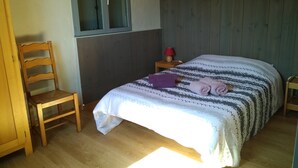 Ett sovrum med en säng i trä, en stol, ett fönster och ett nattduksbord med en lampa.