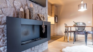 Una sala moderna con una pared de piedra, una chimenea negra, leña y un comedor con mesa y sillas, además de una alfombra de piel blanca.