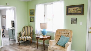 A living room with a striped armchair, a tufted armchair with a blue pillow, a wooden side table with a lamp, and framed artwork on the walls.