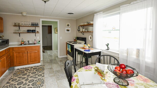 A kitchen with wooden cabinets, a dining table with a floral tablecloth, and a fruit bowl. There is a window with sheer curtains and a view of the outside.