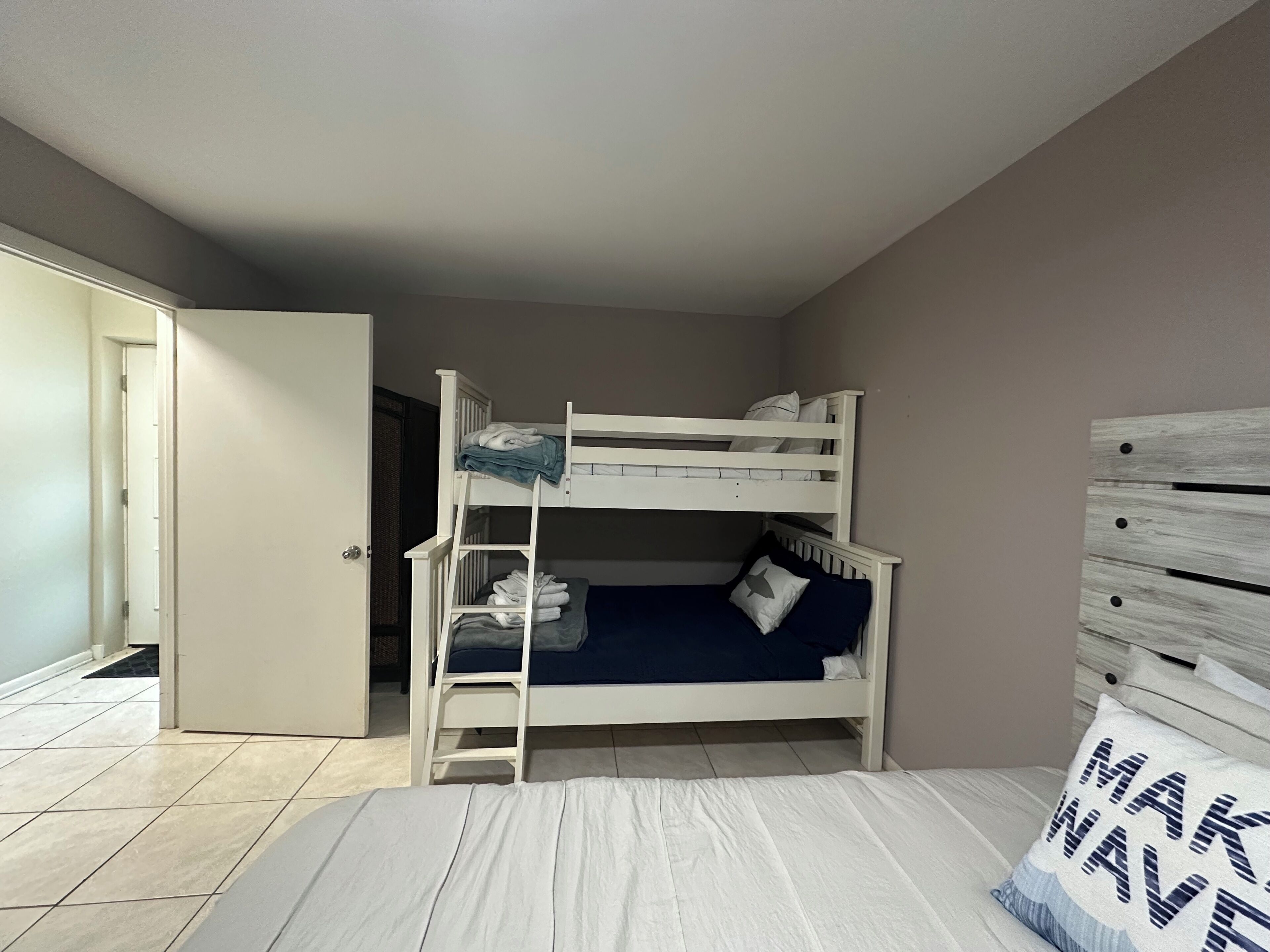 A bunk bed room with a door leading to another room, a bed with a pillow, and a wall with a wooden panel.