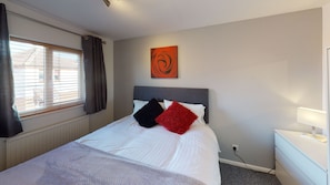 Una cama bien hecha con ropa de cama blanca, dos almohadas en negro y rojo, un cabecero gris y una obra de arte en la pared.