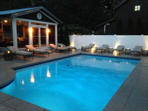 Une piscine rectangulaire bordée de carreaux, entourée de chaises longues et d’un petit bâtiment doté de portes en verre.