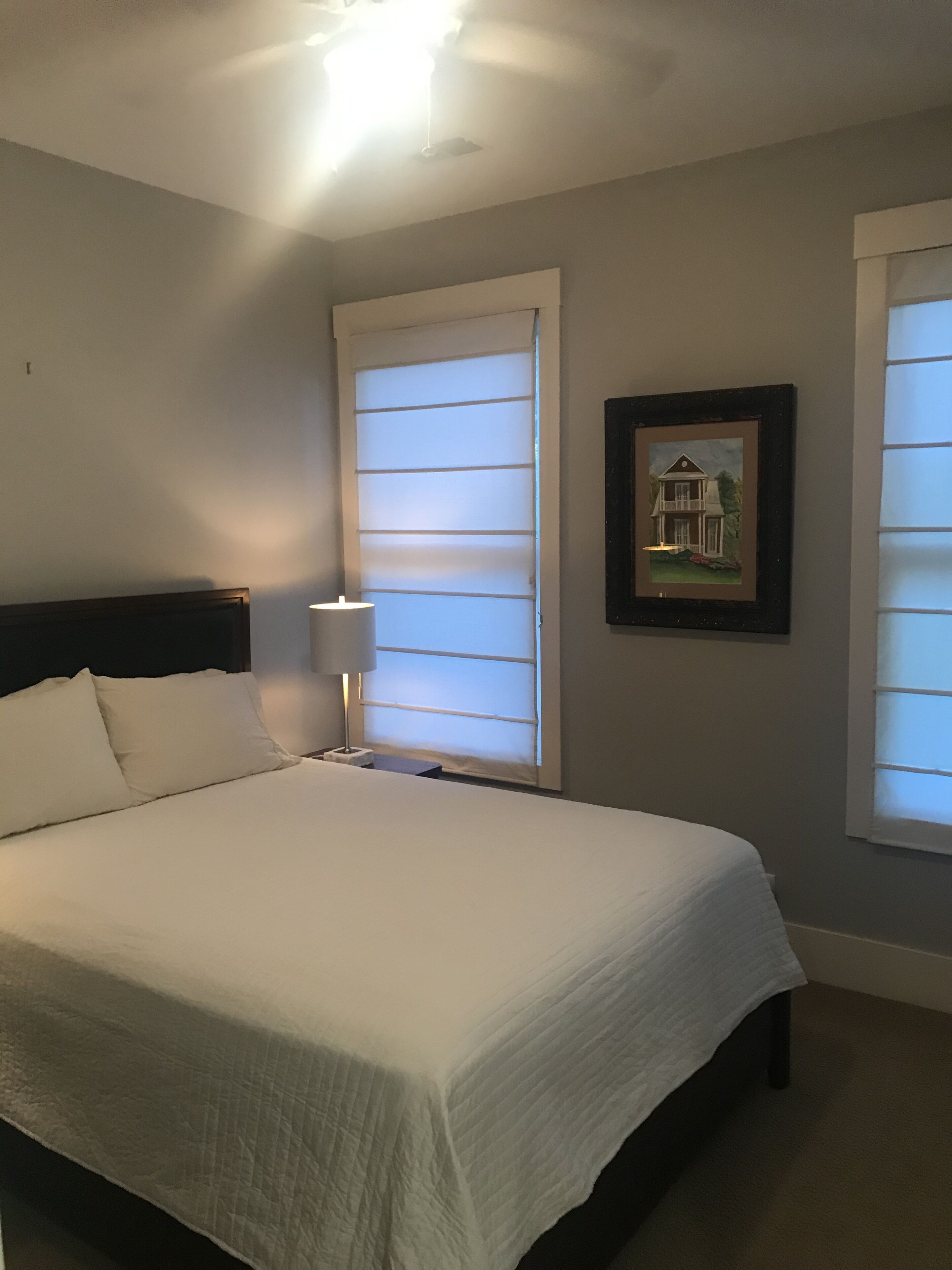 A bedroom with a bed, a nightstand, a lamp, and two windows with blinds.
