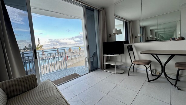 A modern hotel room with a balcony, a TV on a stand, a dining table with chairs, and a large mirror.