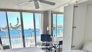 A hotel room with a large bed, a desk, a chair, a TV, and a balcony with a view of the sea and a tower.