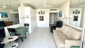 A spacious living room with a sofa, a dining table, and a TV. There are ceiling fans and a door leading to another room.