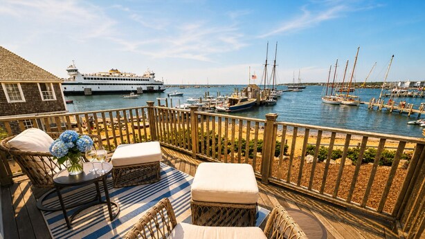 Una terraza de madera con mobiliario de mimbre, una mesa con un jarrón de flores y vistas a un puerto deportivo con barcos.