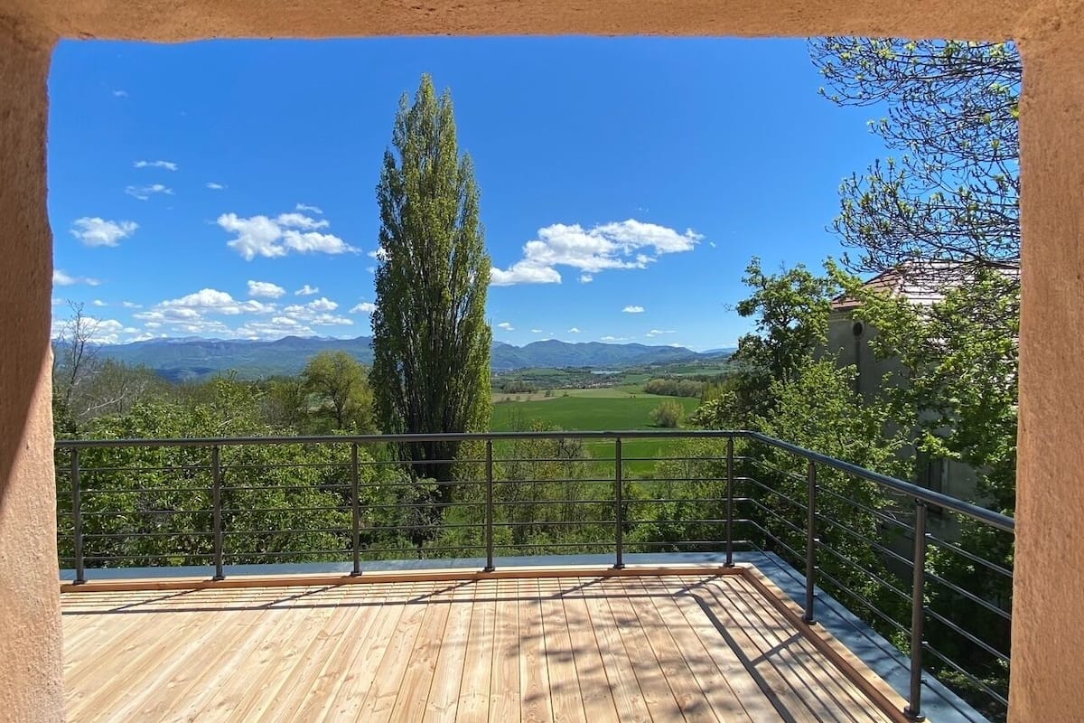 A wooden deck with a metal railing overlooking a green landscape with trees and mountains.
