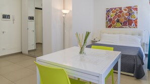 Et hotelværelse med en seng, et natbord, en lampe, et billede på væggen og et bord med papirer samt en glasvase med grene.