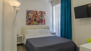 Et hotelværelse med en seng, et natbord, en lampe, et fjernsyn og et farverigt billede på væggen.