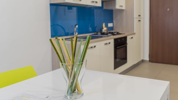 Et moderne køkken med hvide skabe, en blå bagplade og en glassvase med grønne og gule stængler på et hvidt bord.