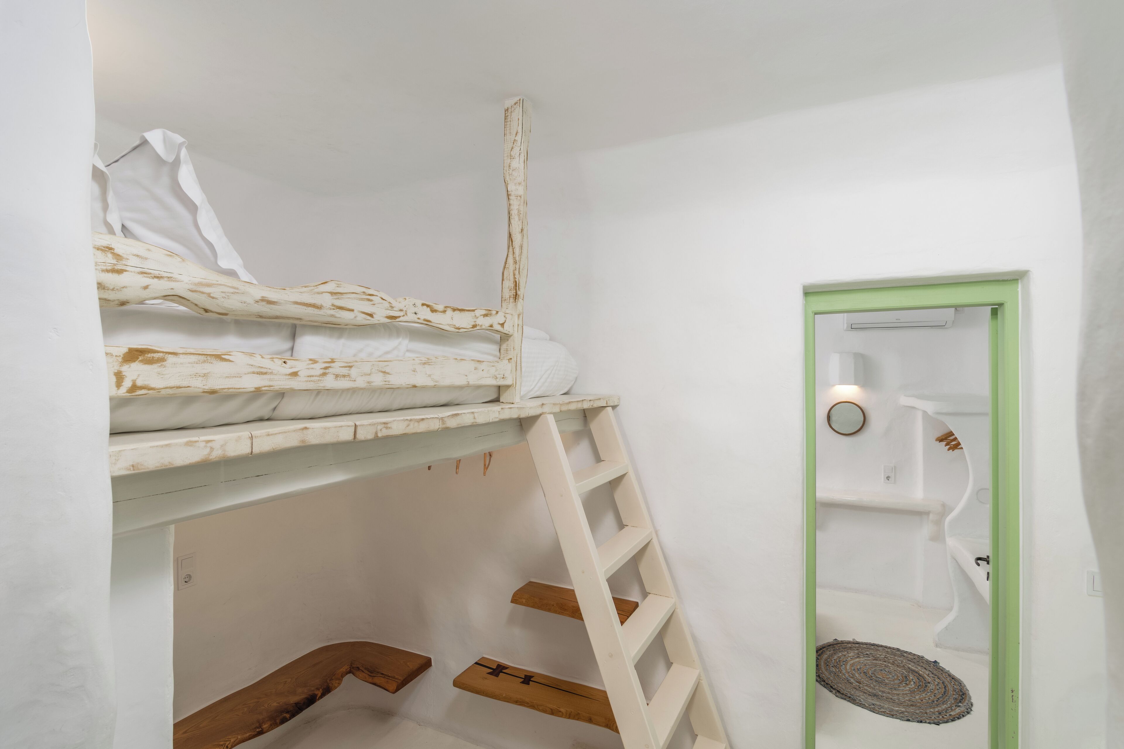 Una cama litera con escalera, un pequeño estante de madera y una puerta con marco verde que da a un baño con toallero y espejo circular.