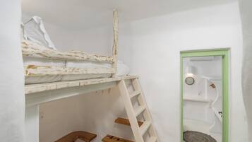 Una cama litera con escalera, un pequeño estante de madera y una puerta con marco verde que da a un baño con toallero y espejo circular.