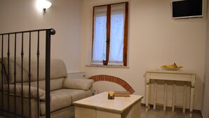 Un saló amb un sofà beix, una taula petita amb una espelma, una taula blanca amb un plat de fruita, un televisor fixat a la paret i una finestra amb cortines.
