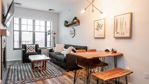 A modern living room with a sofa, coffee table, dining table, and chairs. There is a window with blinds, a floor lamp, and wall-mounted shelves with plants.