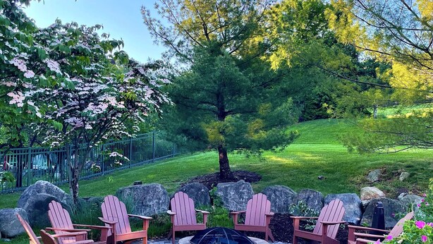 A patio area with Adirondack chairs, a fire pit, and a view of a lush garden and trees.