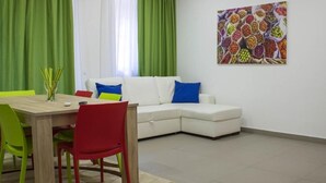 A living room with a white sofa, green curtains, a colorful painting, and a dining area with a wooden table and chairs. - Family three-room apartment (Catania)