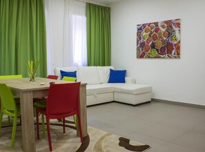 A living room with a white sofa, green curtains, a colorful painting, and a dining area with a wooden table and chairs. - Family three-room apartment (Catania)