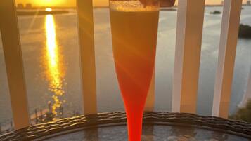 Una copa de cóctel de tonalidad roja, decorada con una hoja de menta y una fresa, sobre una mesa con vista a una puesta de sol sobre el agua.