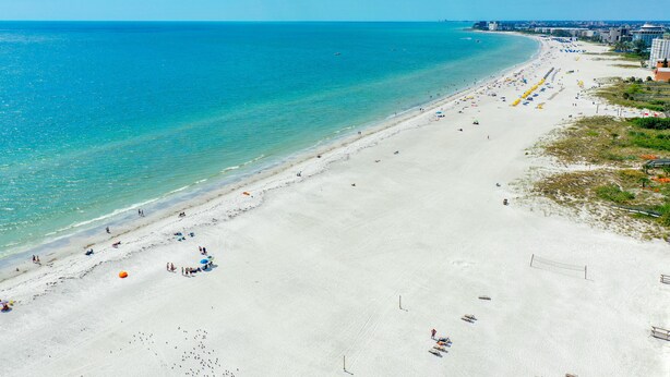 A wide beach with clear turquoise water, a long stretch of sandy shore, and a few buildings in the background.