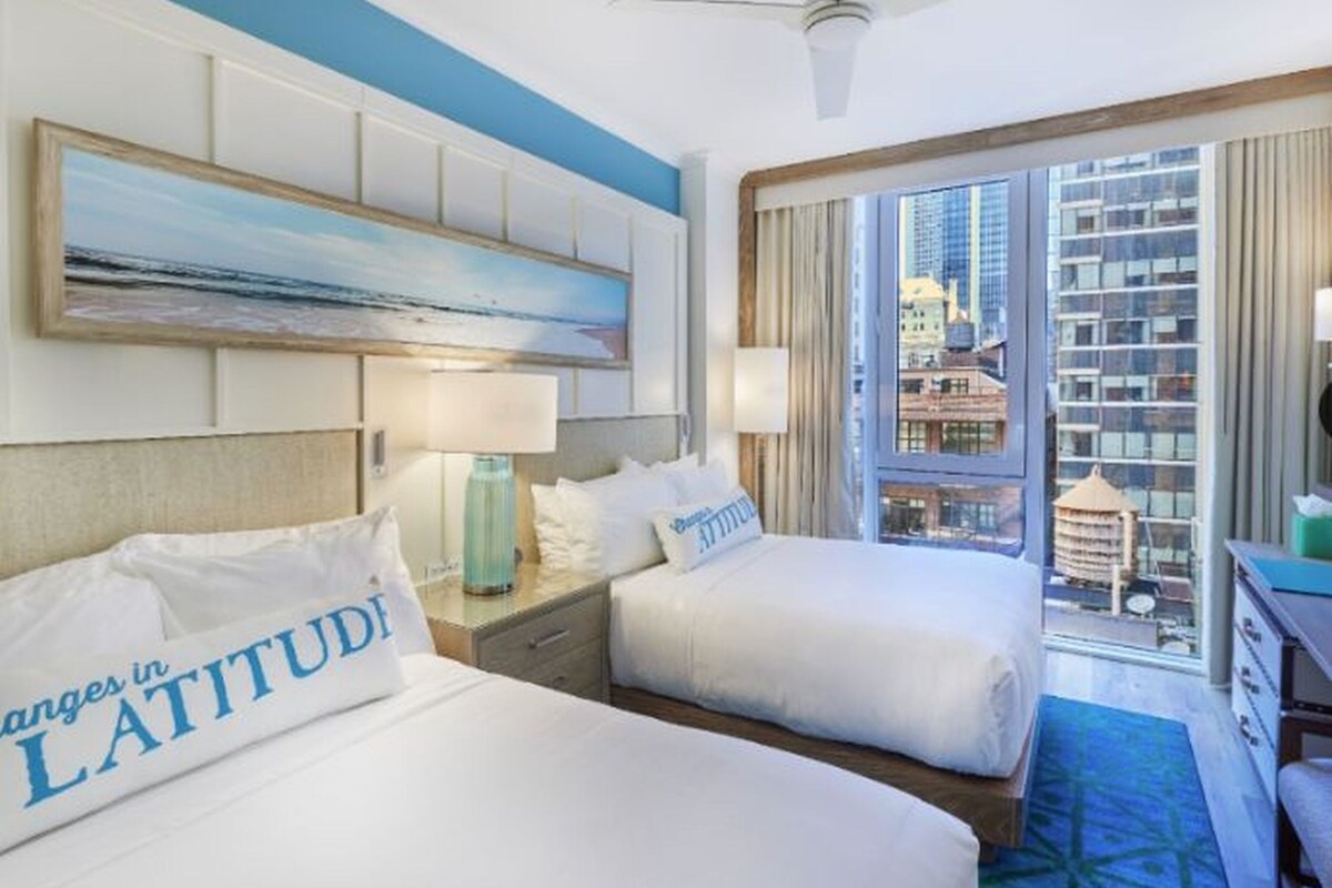 A hotel room with two beds, a ceiling fan, a painting, and a view of the city.