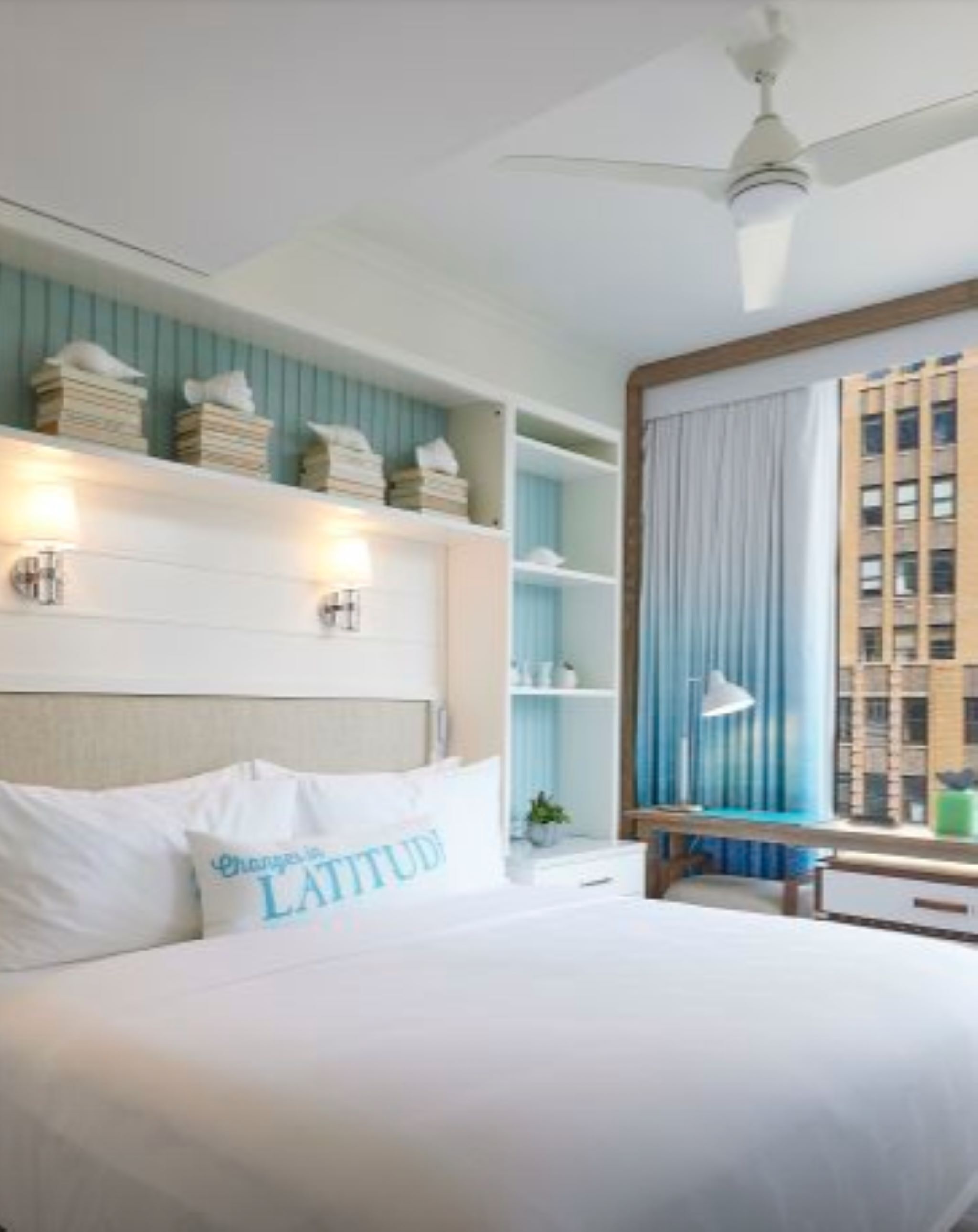 A bedroom with a bed, a ceiling fan, a window with curtains, and a view of a building.
