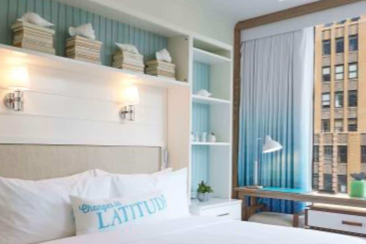 A bedroom with a bed, a ceiling fan, a window with curtains, and a view of a building.