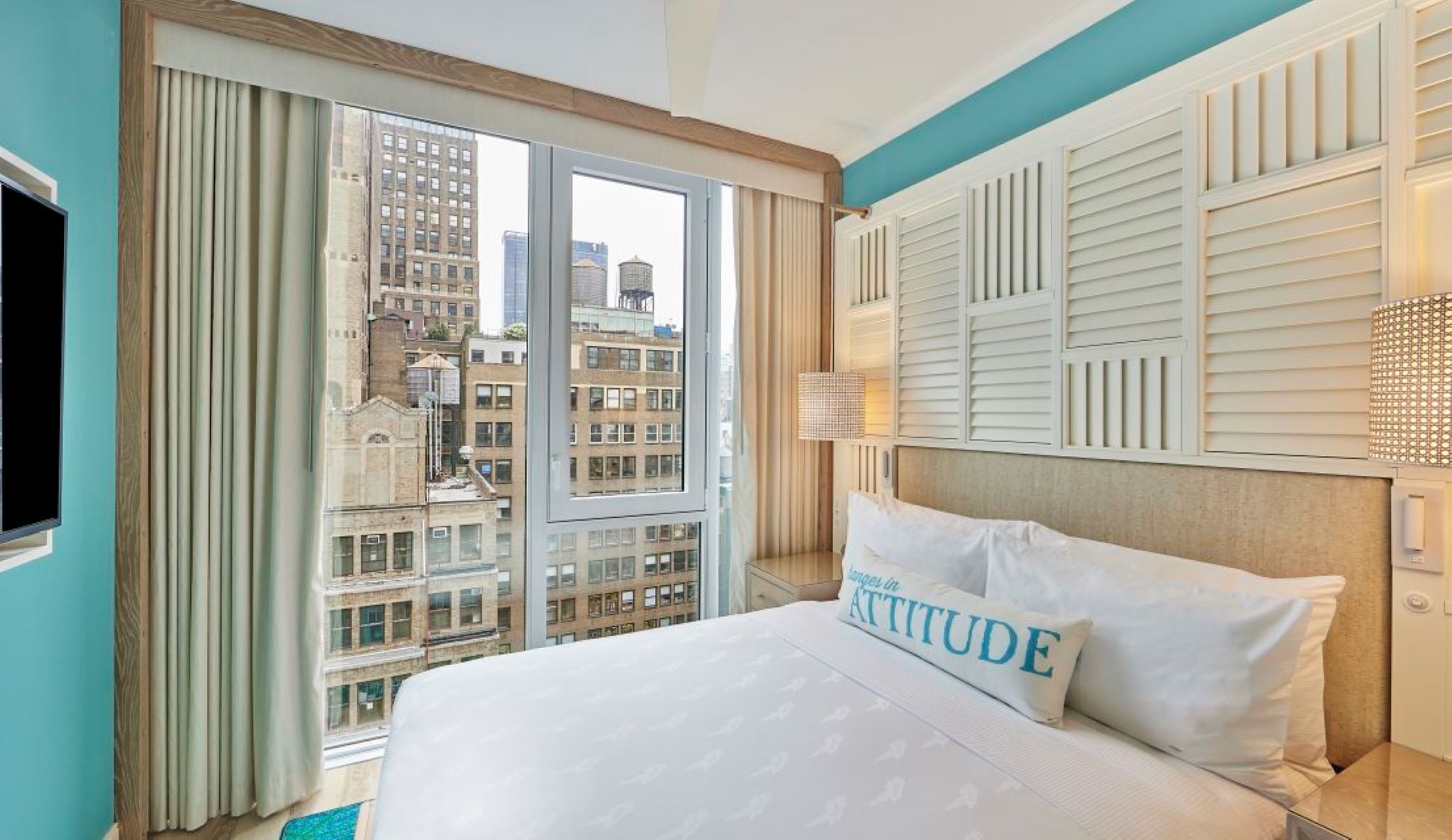 A hotel room with a large bed, a view of city buildings, a TV, and a window with curtains.