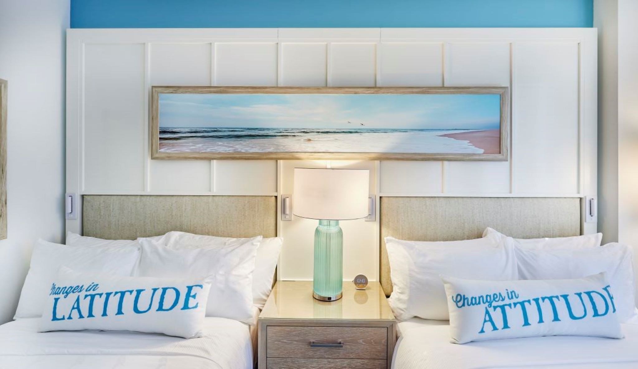 A hotel room with two beds, a nightstand with a lamp, and a painting of a beach scene.