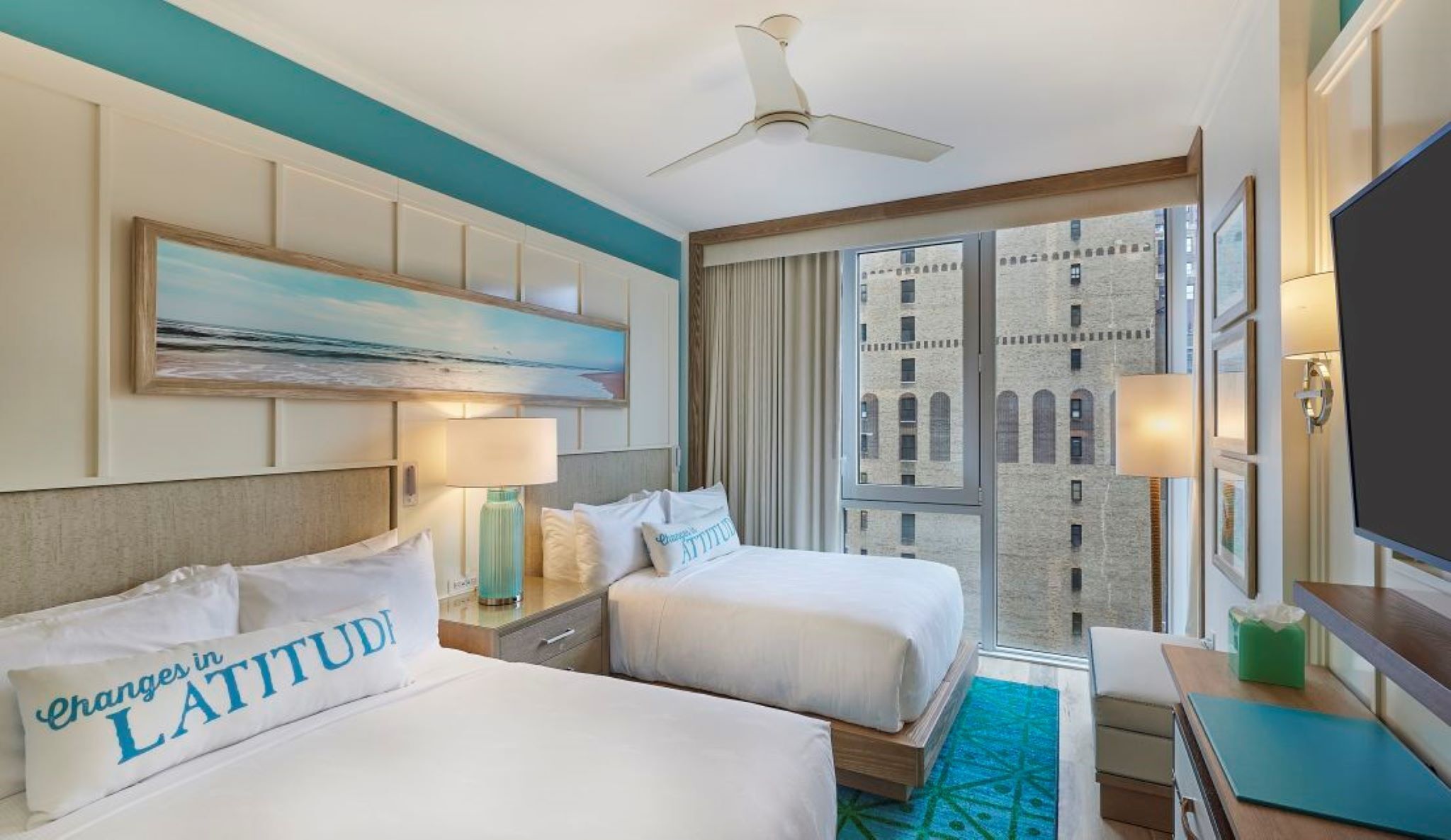A hotel room with two beds, a ceiling fan, a painting, a large window with a view of a building, and a flat-screen TV.