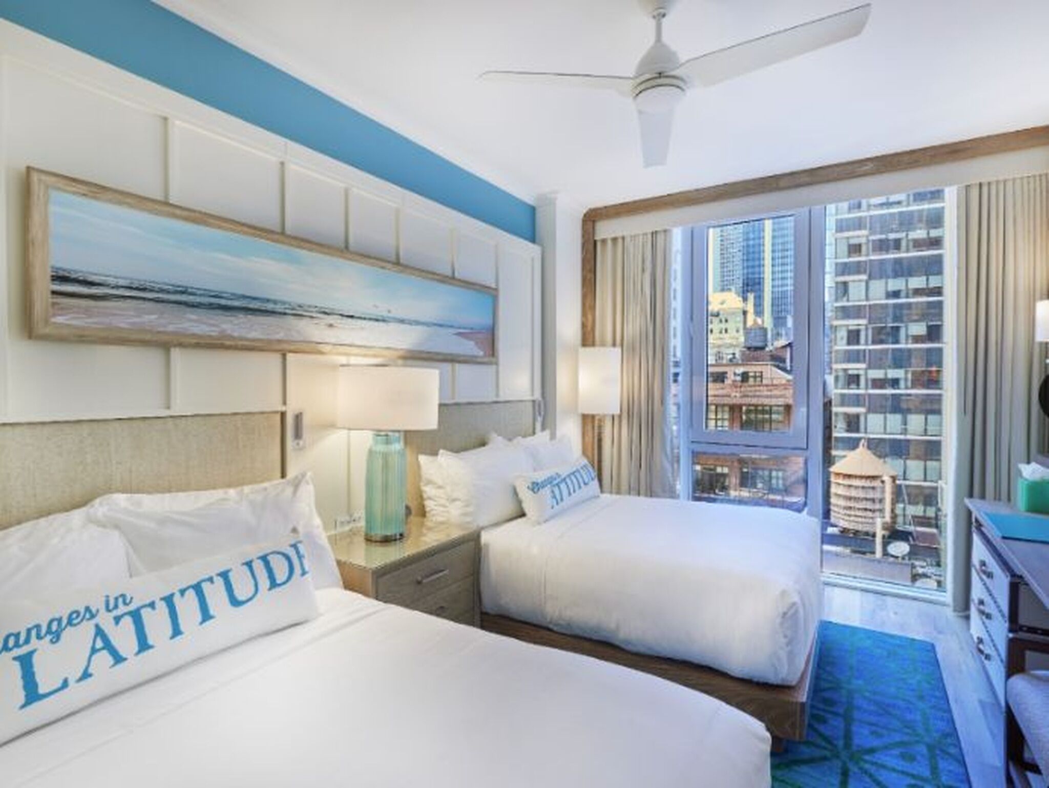 A hotel room with two beds, a ceiling fan, a painting, and a view of the city.