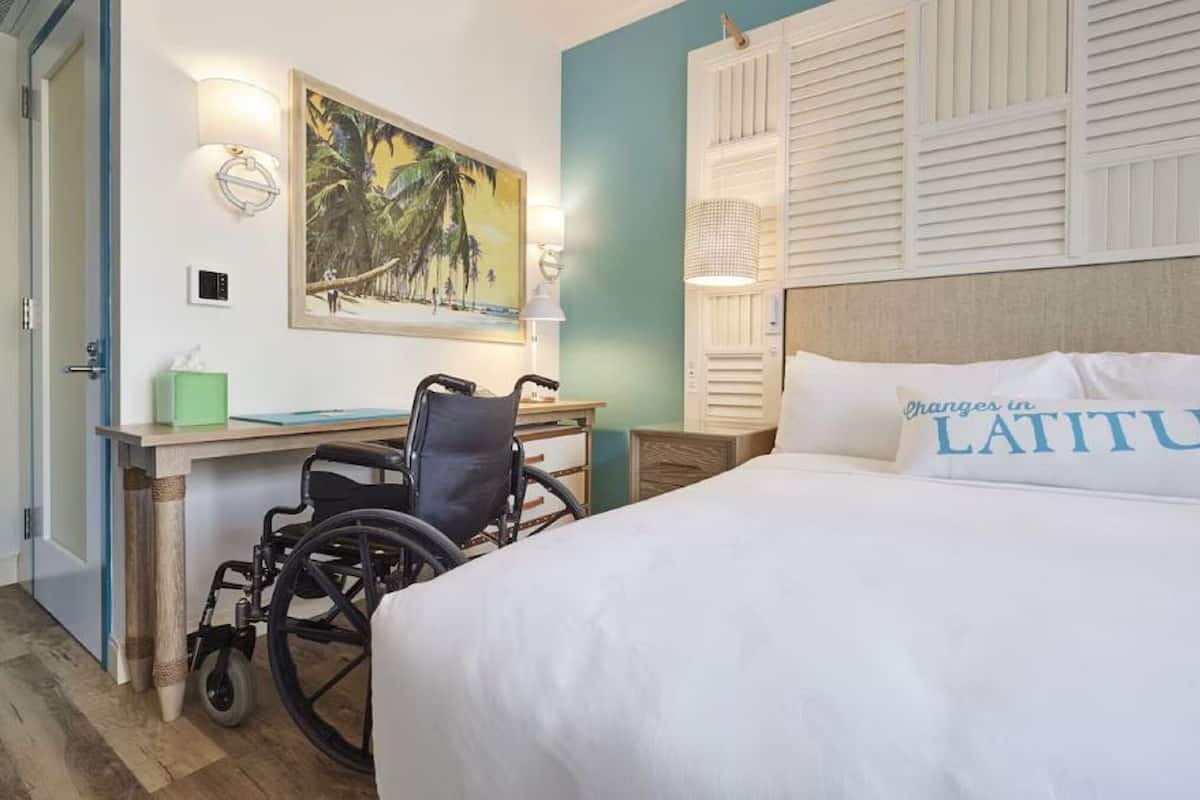 A hotel room with a bed, a desk, a wheelchair, a painting, and a wall-mounted light fixture.