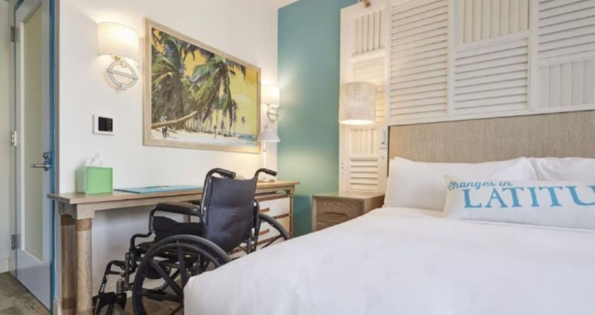 A hotel room with a bed, wheelchair, desk, and a painting on the wall.